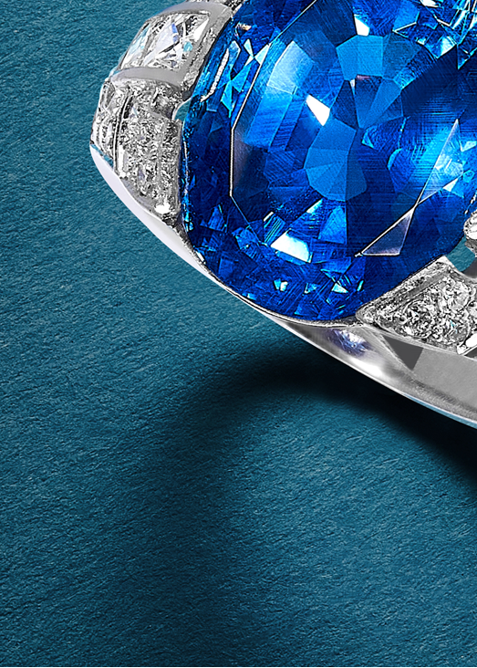 Close up of part of a ring, a faceted blue stone and a square cut clear stone in white metal, on teal