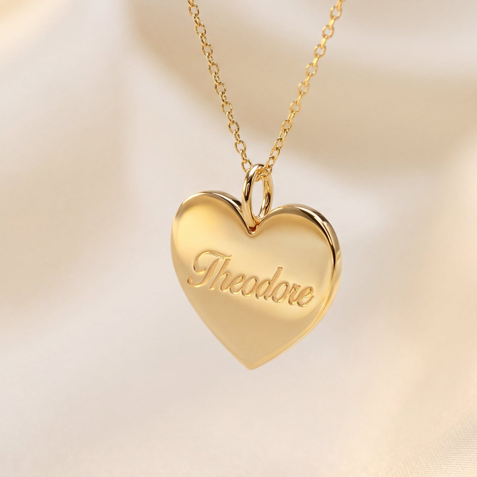 Gold heart-shaped pendant with 'Theodore' engraving on a soft beige background
