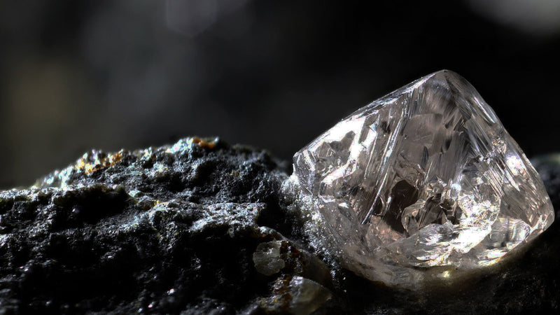 Rough uncut diamond crystal resting on a dark rock, lit from one side against a black background