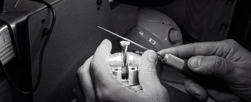 Black and white close up of a jeweller using a fine point on a small stone set ring in a bench clamp