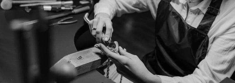 Jeweller in a white shirt and dark apron working at a bench, a silver coloured chain in one hand